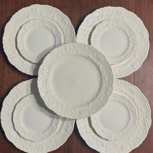 May include: A set of five white ceramic plates featuring a raised, embossed floral pattern along the scalloped edges. The collection includes four larger plates with smaller plates centered on top of them, and one additional plate in the center.