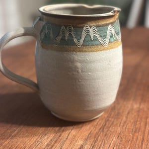 May include: A ceramic pitcher with a speckled off-white base and a handle on the left side. The upper portion features a decorative band with a teal and brown glaze, accented by a series of white, wavy, mountain-like etched lines circling the rim.