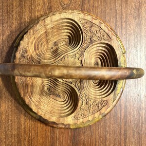 May include: A circular wooden folding basket with a natural brown grain, featuring three carved oval-shaped recessed sections and a central floral motif. The basket includes a horizontal handle spanning across the top surface of the wood.