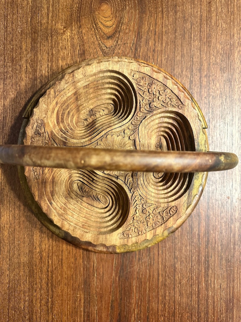 May include: A circular wooden folding basket with a natural brown grain, featuring three carved oval-shaped recessed sections and a central floral motif. The basket includes a horizontal handle spanning across the top surface of the wood.