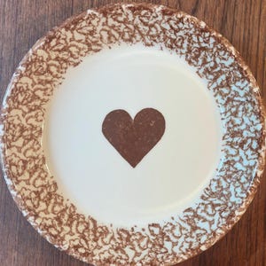 May include: A round ceramic plate with a smooth white center featuring a solid brown heart in the middle. The rim of the plate is decorated with a textured, mottled brown sponge-painted pattern that creates a distinct border around the edge.