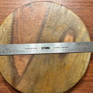 May include: A round wooden board with visible natural grain patterns in shades of brown. A stainless steel C-Thru ruler is placed across the center, showing a diameter of approximately 11.75 inches. The board has a smooth, flat surface.
