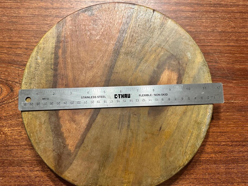 May include: A round wooden board with visible natural grain patterns in shades of brown. A stainless steel C-Thru ruler is placed across the center, showing a diameter of approximately 11.75 inches. The board has a smooth, flat surface.