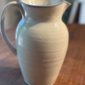 May include: A cream-colored ceramic pitcher with a speckled finish and a smooth, glossy glaze. The vessel features a rounded body, a wide pouring spout, and a curved handle on the side. The rim is accented with a subtle, thin band of light blue.