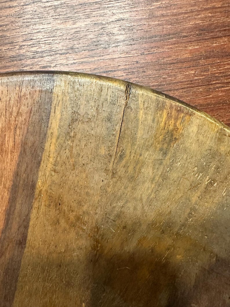 May include: A circular wooden board featuring a natural grain pattern with varying shades of brown, ranging from light tan to deep mahogany. A thin, vertical crack is visible near the curved edge of the board, extending toward the center.