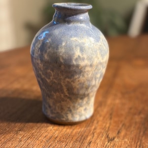 May include: A small ceramic vase with a rounded body and a narrow neck. The surface features a mottled glaze with blended shades of periwinkle blue and creamy off-white, creating a textured appearance. The rim is smooth and slightly flared outward.
