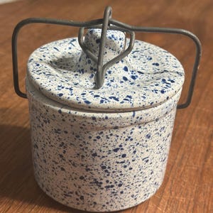 May include: A cylindrical ceramic canister with a white base and blue speckled pattern. The canister features a matching lid with a central knob and a gray metal wire bail handle that arches over the top to secure the lid in place.
