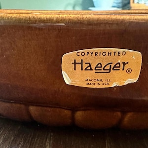 May include: A brown glazed ceramic bowl with a scalloped base and a tan sticker that reads COPYRIGHTED Haeger MACOMB, ILL. MADE IN U.S.A. The bowl features a smooth, glossy finish and a rounded rim, showcasing a deep, warm brown color throughout.