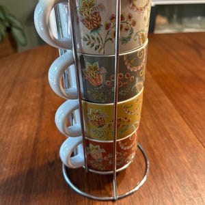 May include: A set of four stacked ceramic mugs held in a vertical chrome metal stand. Each mug features a white handle and a colorful floral pattern with leaves and vines. The mugs are colored in white, gray, mustard yellow, and burnt orange.