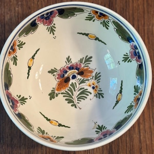 May include: A round ceramic bowl with a cream-colored base featuring hand-painted floral motifs in shades of orange, pink, and green. The interior rim is decorated with a thin blue line, and the center displays a detailed floral arrangement.