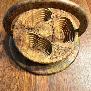 May include: A circular wooden serving tray featuring a carved floral center and three recessed, oval-shaped compartments. The tray includes a large, arched handle with decorative engravings and a scalloped edge, showcasing natural wood grain patterns.