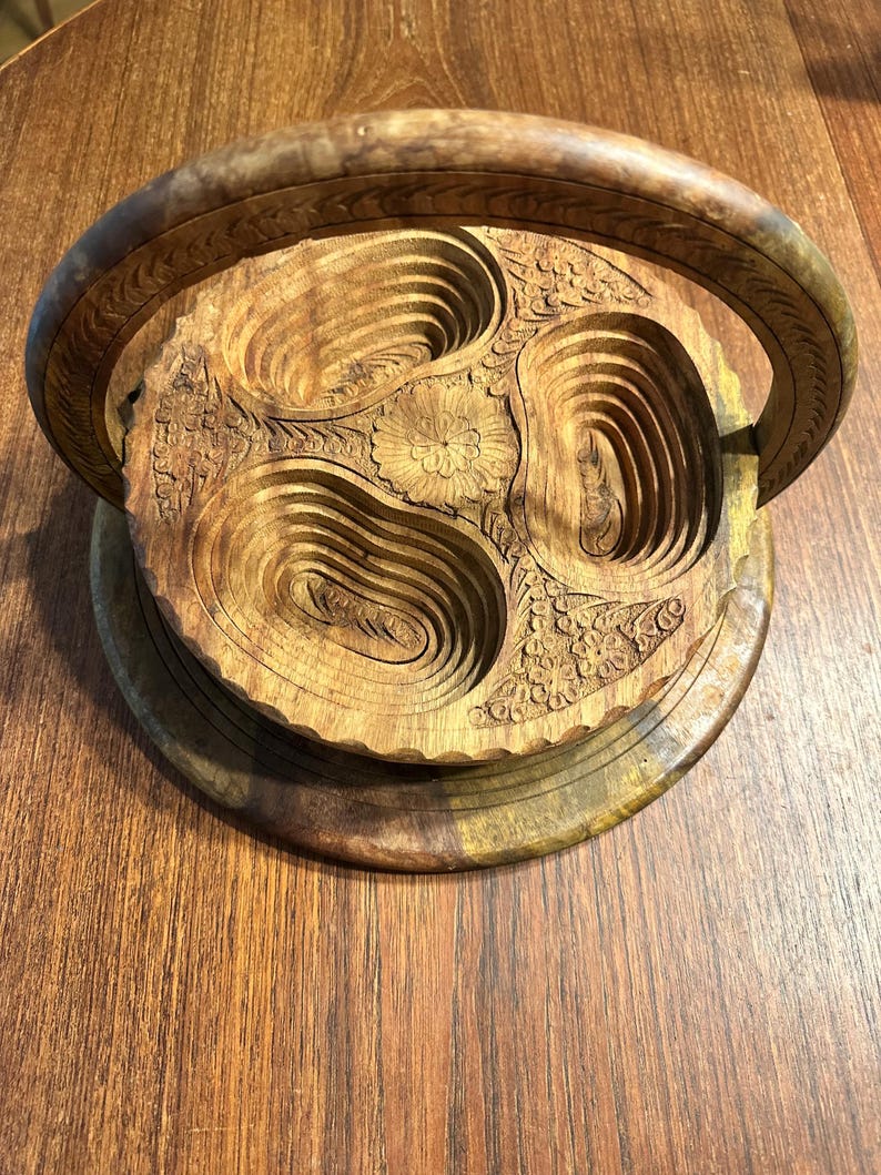 May include: A circular wooden serving tray featuring a carved floral center and three recessed, oval-shaped compartments. The tray includes a large, arched handle with decorative engravings and a scalloped edge, showcasing natural wood grain patterns.