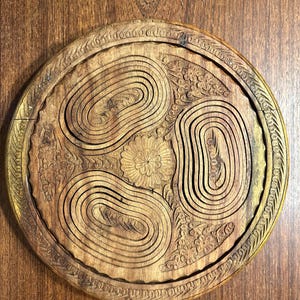 May include: A circular wooden trivet featuring a carved floral center surrounded by three concentric oval patterns. The outer edge is decorated with a repeating leaf motif. The wood displays a natural brown grain with visible carved textures.