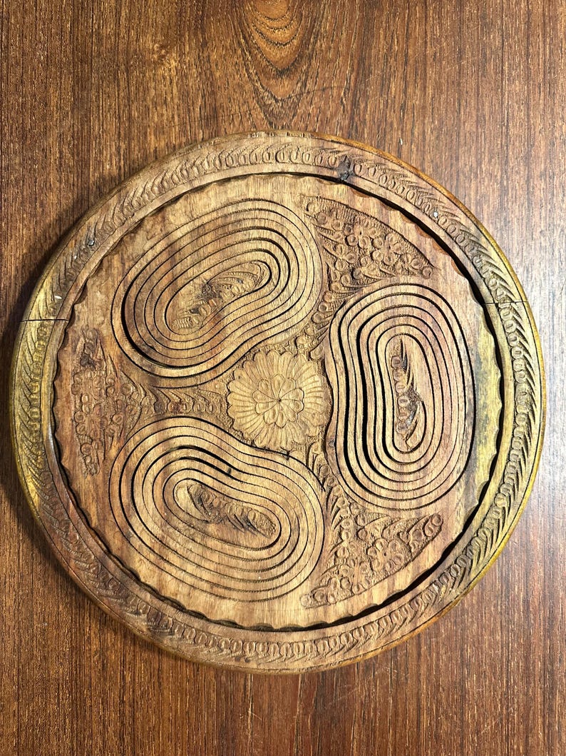 May include: A circular wooden trivet featuring a carved floral center surrounded by three concentric oval patterns. The outer edge is decorated with a repeating leaf motif. The wood displays a natural brown grain with visible carved textures.