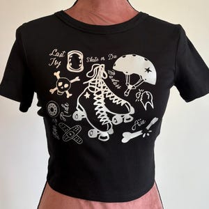Può includere: Top corto nero con grafica bianca a tema pattinaggio a rotelle. Il design include un pattino, un casco e il testo "Last Try", "Skate or Die", "Badass" e "Aie".