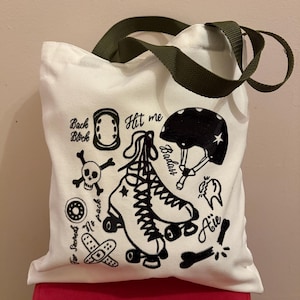 Peut inclure: Sac fourre-tout en toile blanche avec des poignées vert olive. Il présente des illustrations noires de patins à roulettes, d'un casque et d'autres graphiques liés au patinage. Le texte comprend "Hit me" et "Badass".