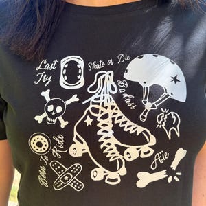 Peut inclure: T-shirt noir avec un motif blanc représentant des patins à roulettes, un casque, des genouillères, une tête de mort et d'autres éléments liés au patinage. Le texte comprend "Skate or Die", "Last Try" et "Born to Ride".
