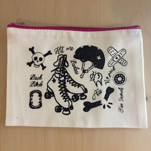 Peut inclure: Une pochette rectangulaire en toile avec une fermeture éclair rose et des illustrations noires. Les illustrations comprennent une tête de mort, des patins à roulettes, un casque, des pansements et du texte.
