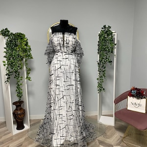 May include: A full-length, off-the-shoulder gown on a mannequin. The white fabric is patterned with black lines. The dress has a fitted bodice and a flowing skirt. The mannequin is on a gold-coloured circular base.