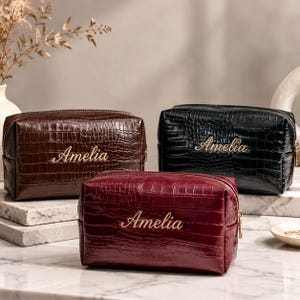 Può includere: Tre pochette rettangolari in marrone scuro, nero e rosso intenso, ciascuna con una texture effetto coccodrillo e il nome Amelia ricamato in filo dorato sul davanti, con una chiusura a zip color oro sulla parte superiore.