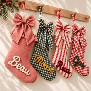 May include: Four Christmas stockings hanging on a wooden rack. The first is solid dusty rose with a large bow and the name Beau. The second is green and white gingham with a bow and the name Moose. The third is red striped with a bow and the name Rua. The fourth is red and white gingham with a bow and the name Gus.