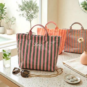 Puede incluir: Tres bolsos tote con rayas verticales rosas, naranjas y grises. El bolso delantero tiene rayas verdes y el nombre "Lola" en cursiva. Gafas de sol con una cadena dorada descansan sobre la mesa. También hay un vaso de menta y un jarrón pequeño.
