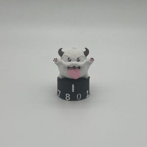 May include: A small white creature figurine with two brown horns, pink paw pads, and a protruding pink tongue. The creature sits atop a black cylindrical base featuring a vertical white line and the white numbers 7, 8, 0, and 1 printed around it.