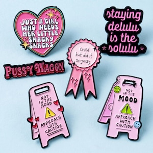 May include: A collection of six enamel pins featuring various designs: a heart with text, a ribbon award with text, a horizontal bar with text, and two caution signs with text. The pins are colored in shades of pink, black, yellow, and white.
