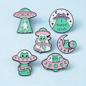 Puede incluir: Un conjunto de seis pines de esmalte con gatos y ovnis en tonos rosa pastel y verde menta. Los diseños incluyen gatos en platillos voladores, un gato en una luna creciente y los textos I WANNA LEAVE, SET ME-OWT OF HERE y TAKE ME con estrellas.