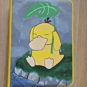 May include: A yellow and gray card binder featuring a cartoon duck holding a leaf umbrella in the rain. The binder has a yellow border and the text "V-itx" is visible on the bottom left corner. The background is a gray sky and green grass.