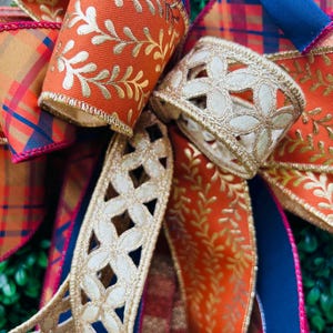 May include: A decorative bow made of multiple ribbons featuring orange fabric with a gold metallic leaf pattern, a navy blue and orange plaid pattern, and a cream-colored ribbon with a laser-cut floral design and gold metallic edges.