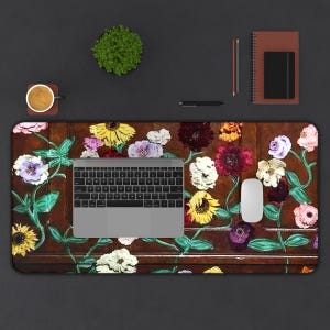 May include: A floral-patterned desk mat with a laptop, mouse, and keyboard. The mat features a brown background with colorful flowers and green vines. A cup of coffee, a notebook, and a pen are also on the dark surface.