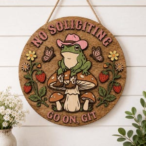 Puede incluir: Un letrero redondo de madera con cuerda para colgar que muestra una rana verde con sombrero de vaquero rosa y botas marrones sentada sobre un hongo. El texto dice NO SOLICITING y GO ON, GIT. Incluye mariposas rosas, flores amarillas y fresas rojas.