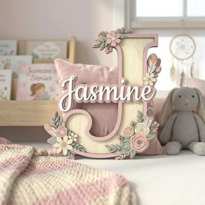 Könnte beinhalten: Ein Holzbuchstabe "J" mit dem Namen "Jasmine" in Schreibschrift. Der Buchstabe ist mit floralen Akzenten in Rosa, Weiß und Grün verziert. Ein rosa Kissen und ein graues Stoffkaninchen befinden sich im Hintergrund. Das Gesamtthema ist Kinderzimmerdekoration.