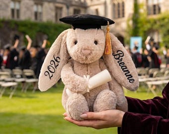 May include: A plush bunny wearing a black graduation cap with a gold tassel, holding a rolled diploma. The bunny's ears are embroidered with "2026" and "Breanne". The bunny is light brown and is held in front of a graduation ceremony.