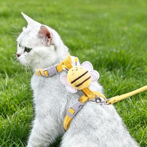 May include: A yellow cat harness with a bee-shaped decoration. The harness has a matching collar and leash, all in yellow. The bee decoration is yellow and brown with white accents. The cat is white and sitting on green grass.