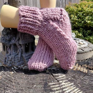 May include: A pair of chunky knit socks in a dusty mauve color. The socks feature a thick ribbed cuff at the top and a standard stockinette stitch pattern throughout the foot and heel area, crafted from a soft, textured yarn with visible loops.
