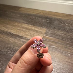 May include: A silver-toned earring featuring a flower design with pink and clear crystals. The earring has a small, dangling flower at the bottom. The earring is held up by a person's hand.