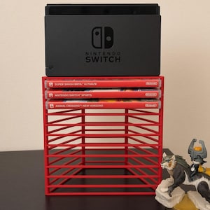 May include: A black Nintendo Switch console dock sits atop a red plastic storage rack containing three game cases: Super Smash Bros. Ultimate, Nintendo Switch Sports, and Animal Crossing: New Horizons. A wolf-like figurine stands to the right.