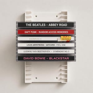May include: A white plastic wall-mounted rack holding six music cassette cases stacked horizontally. The spines display titles including The Beatles Abbey Road, Daft Punk Random Access Memories, OneRepublic, Louis Armstrong Satchmo, Beethoven Symphony No. 5, and David Bowie Blackstar.
