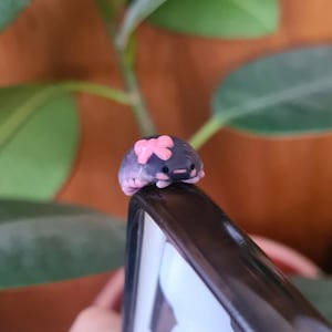 May include: A small, sculpted creature figurine with a dark purple, rounded body and a light pink bow on its head. The figure has tiny pink accents on its sides and a simple face with two small black eyes and a horizontal pink line for a mouth.