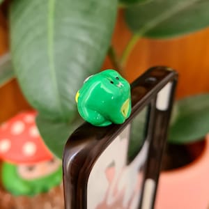 May include: A small, glossy, bright green frog figurine with a smooth surface, two black dots for eyes, and a yellow accent on its side, perched on the top edge of a dark-framed smartphone case with a white and pink patterned insert.