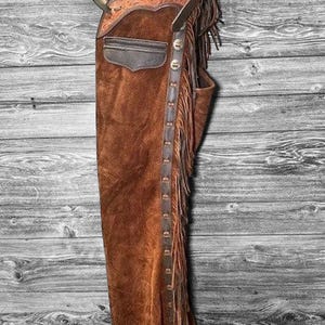 Puede incluir: Chaps de cuero de gamuza marrón con repujado decorativo en la parte superior. Los chaps tienen un bolsillo, flecos a los lados y una serie de broches metálicos en la parte delantera. Los chaps cuelgan sobre un fondo de madera envejecida.