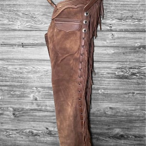 May include: Brown suede leather chaps with fringe detailing. The chaps feature a pocket, decorative stitching, and silver-tone button accents. They are hanging against a rustic wooden background.