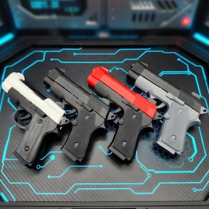 Puede incluir: Cuatro pistolas de juguete en fila, cada una con un esquema de color diferente. La primera es blanca y negra, la segunda y tercera son negras con la parte superior negra o roja, y la cuarta es gris y negra. Están dispuestas sobre una superficie negra con líneas azules de neón.