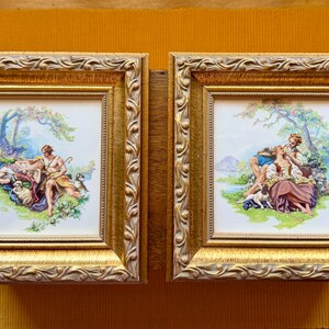 May include: Two square wall plaques featuring ornate gold-toned frames with a decorative leaf-like border. Each plaque contains a central ceramic tile depicting a colorful pastoral scene with figures, trees, and animals in a grassy landscape setting.