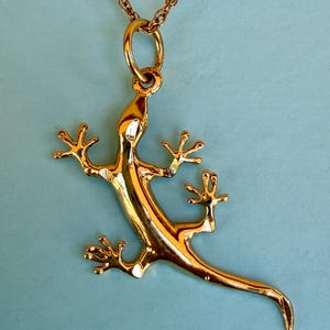 May include: A gold-colored lizard pendant necklace. The lizard is detailed with outstretched limbs and a long tail. The necklace chain is also gold-colored, and the pendant hangs against a light blue background. The jewelry is likely made of metal.