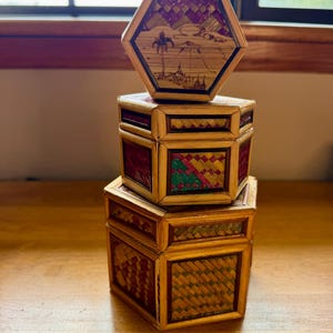 May include: A stack of three hexagonal boxes crafted from woven straw and reed. The top box features a landscape illustration, while the middle and bottom boxes display geometric patterns in shades of red, green, yellow, and natural tan.