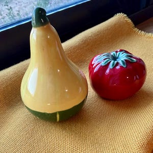 May include: A yellow pear-shaped ceramic figurine with a green stem and base, next to a red tomato-shaped ceramic figurine with a green top. Both figurines are on a yellow cloth.