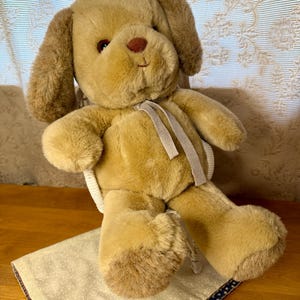 Puede incluir: Un peluche de cachorro de color canela con orejas caídas, una nariz marrón y ojos oscuros. El perro está sentado sobre una tela estampada con un borde rojo y azul. Una cinta de color claro está alrededor del cuello del perro.
